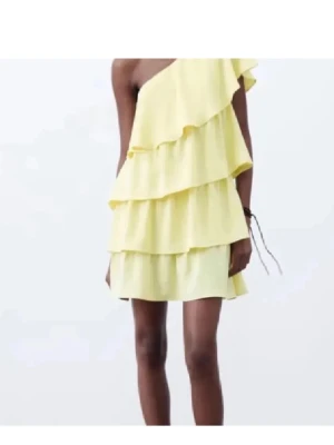 Gul one shoulder volangklänning - Superfin gul kortklänning med flera volanger och oneshoulder-design. Klänningen har en luftig och lätt känsla, perfekt för varma dagar. Den är ärmlös på ena sidan och har en snygg asymmetrisk look som sticker ut.