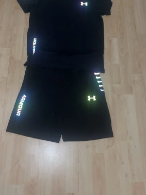 Svart träningsset från Under Armour - Snyggt svart träningsset från Under Armour med t-shirt och shorts. Setet har reflekterande loggor och textdetaljer i vitt och regnbågsskimrande färg. Perfekt för träning och sportiga aktiviteter. Materialet är lätt och andas, vilket gör det bekvämt under intensiva pass.