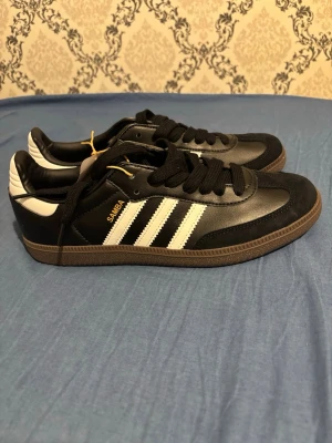 Adidas Samba svartvita sneakers - Klassiska Adidas Samba sneakers i svart och vitt med de ikoniska tre ränderna på sidan. Skorna har ovandel i läder och mocka, gummisula och blå Adidas-logga på plösen. Perfekta för dig som gillar retrostil och streetwear.