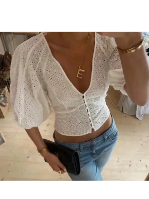 Vit broderad blus  - Superfin vit blus med broderade detaljer och v-ringning. Toppen har puffiga trekvartsärmar, knappar framtill och är croppad i modellen. Perfekt för dig som gillar romantisk och trendig stil.