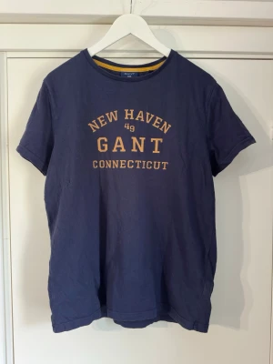 Blå GANT t-shirt med tryck - Mörkblå t-shirt från GANT med gul text 'NEW HAVEN GANT CONNECTICUT' på bröstet. Klassisk rund halsringning och korta ärmar. Perfekt för en avslappnad stil.