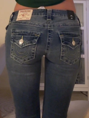 Blå True Religion jeans med fickdetaljer - Säljer ett par blå jeans från True Religion med ikoniska kontrastsömmar och snygga ficklock bak med silverfärgade knappar. Jeansen har en klassisk femficksmodell och är tillverkade i denim med lätt tvättad look. Perfekta för dig som gillar streetstyle och detaljer.