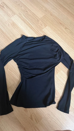 Svart långärmad topp från Gina Tricot - Svart långärmad topp från Gina Tricot med asymmetrisk rynkning på ena sidan. Tunn och stretchig modell med rund halsringning, perfekt för en trendig och stilren look.