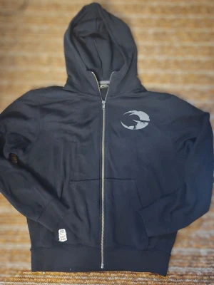 Svart zip-hoodie från GASP - Svart hoodie med dragkedja från GASP. Stor logga i grått och orange på ryggen samt mindre logga på bröstet. Utrustad med huva och kängurufickor. Perfekt för dig som gillar sportig och avslappnad stil.