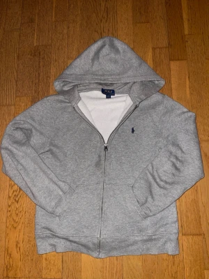 Grå hoodie från Polo Ralph Lauren - Snygg grå zip-hoodie från Polo Ralph Lauren med klassisk blå logga broderad på bröstet. Hoodien har dragkedja, huva och kängurufickor framtill. Mjuk insida i bomullsblandning som gör den extra skön att ha på sig. Det står 14-16 på storleken men passar nog mer en 14 åring pågrund av att den krympt i tvätten.