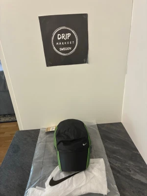 Svart Nike keps med gröna detaljer - Snygg svart keps från Nike med gröna sömmar längs sidorna och klassisk böjd skärm. Kepsen har en liten vit Nike-logga framtill och är tillverkad i ett lätt och sportigt material. Perfekt för en streetwear-look eller till träningen.
