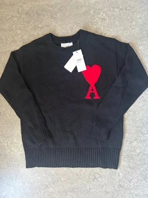 Svart stickad tröja från Ami Paris - Svart stickad tröja från Ami Paris med röd hjärtlogga och bokstaven A på bröstet. Tröjan har rund hals, ribbade muddar och nederkant. Perfekt för dig som gillar stilrena plagg med en twist.