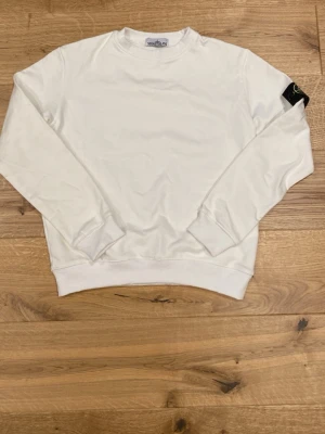 Vit sweatshirt från Stone Island - Säljer en stilren vit sweatshirt från Stone Island med klassisk rund hals och ribbade muddar. Tröjan har den ikoniska Stone Island-loggan på vänster ärm. Perfekt för en clean och modern look.