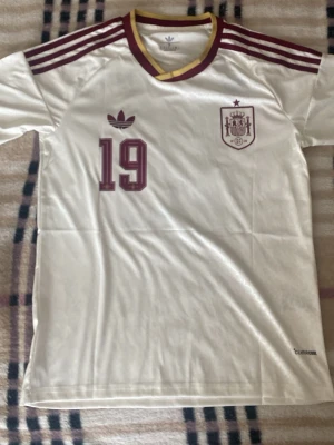 Spanien fotbollströja Adidas #19 - Vit Spanien fotbollströja från Adidas med vinröda detaljer, nummer 19 och namntryck Lamine Yamal på ryggen. Tröjan har klassiska Adidas-ränder på axlarna, broderad landslagssköld och gul/vinröd krage. Tillverkad i lätt och ventilerande material. Helt ny med tag. Priset ej hugget i sten!