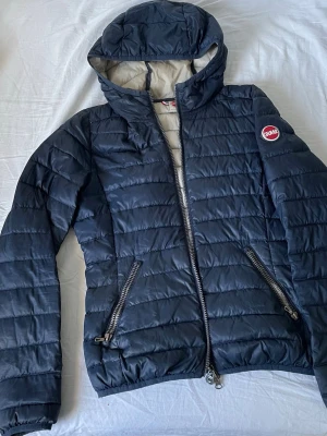Mörkblå dunjacka från Colmar - Snygg mörkblå dunjacka från Colmar med huva och två dragkedjeförsedda fickor. Jackan har quiltad design och Colmar-logga på bröstet. Perfekt för kyliga dagar och enkel att matcha med olika outfits.