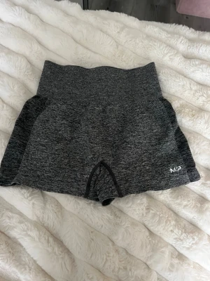 Grå träningstights-shorts från MP - Mörkgrå, tighta träningstights-shorts från MP med hög midja och diskret logga framtill och baktill. Tillverkade i stretchigt syntetmaterial som passar perfekt för gym eller annan träning. Snygg melerad look och korta ben. Passar xs och s 