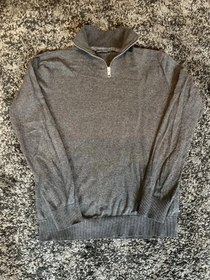 Grå halvzip tröja i bomull från Jack&Jones  - Mörkgrå tröja med halv dragkedja och hög krage. Tillverkad i mjuk bomullsmix med ribbade muddar vid ärmslut och nederkant. Perfekt för lager på lager och enkel att matcha med olika outfits.