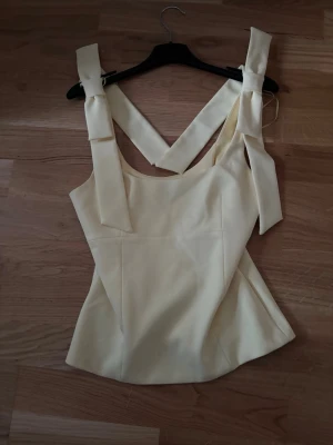 Zara Topp/blus - Ljus gul blus från Zara med breda axelband och stora rosetter på axlarna. Toppen är figursydd och är ärmlös, perfekt inför sommaren. Den har även lätt utsvängd nederkant. Använd 2 gånger och i utmärkt skick 