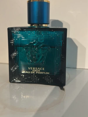 Versace Eros Eau de Parfum - Versace Eros Eau de Parfum i en stilren, fyrkantig flaska i blågrönt glas med guldtext och ikonisk Medusa-detalj. 50 ml med cirka två tredelar kvar. Flaskan har ett blått och guldigt lock som ger en lyxig känsla. Perfekt för dig som gillar exklusiva dofter och snygg design.