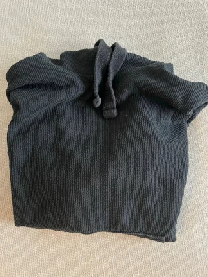 Svart ribbad hoodie med ficka - Snygg svart hoodie med ribbad struktur i storlek M med ficka framtill. Hoodien har luva och snören.