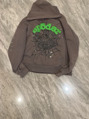 Brun Spider hoodie med tryck - Cool brun hoodie från Spider med stor grön text och spindelnätsmönster på ryggen. Hoodien har huva och detaljerade tryck på ärmarna. Tillverkad i bomull och perfekt för dig som gillar streetwear vibes.