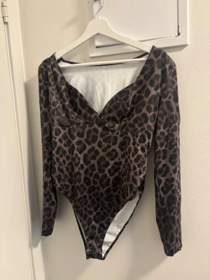 Leopardmönstrad body med lång ärm - Snygg body med leopardmönster och långa ärmar. Djup v-ringning framtill och åtsittande passform. Materialet ser ut att vara stretchigt och mjukt, perfekt för en trendig look. Ingen synlig logga eller märke.