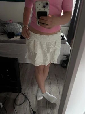Vit volangkjol med smock i midjan - Supersöt vit kortkjol med volanger och smockad midja. Kjolen har ett strukturerat tyg och är perfekt för dig som gillar en luftig och trendig stil. Passar till både sneakers och sandaler för en fräsch look.