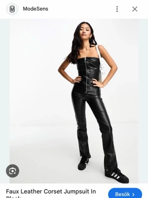 Svart jumpsuit i fuskläder med dragkedja - Snygg svart jumpsuit i fuskläder med figurnära passform och trendig dragkedja framtill. Axelbandslös design med korsettliknande överdel som ger en edgy look. Perfekt för dig som vill sticka ut och köra på en cool stil.