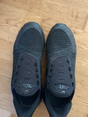 Svarta Nike Air Max 270 sneakers - Snygga helsvarta Nike Air Max 270 sneakers med mesh-överdel och tydlig Air Max-dämpning i hälen. Skorna har en sportig design utan klassiska skosnören och diskreta Nike-loggor på sidan och plösen. Perfekta för en streetwear-look. Skosnören går att få till om man vill. 