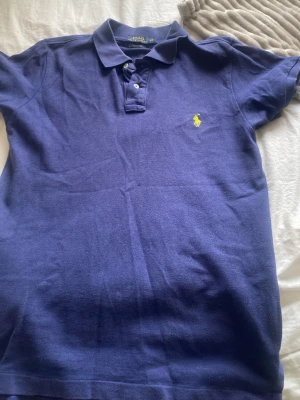 Piké från Polo Ralph Lauren inköpspris 900kr (aldrig använd)  - Klassisk marinblå pikétröja från Ralph Lauren med gul broderad logga på bröstet. Tröjan har krage, knappar framtill och korta ärmar. Tillverkad i bomull för en bekväm och snygg look.