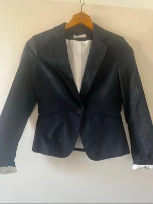 Svart kavaj med klassisk passform - Stilren svart kavaj med en knapp framtill och två fickor. Kavajen har klassisk krage och vit innerfoder som syns vid ärmslut. Perfekt för en dressad look och passar till många olika outfits. Linnemix 