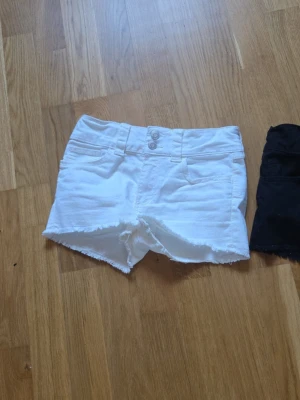 Vita och svarta jeansshorts - Två par klassiska jeansshorts, ett par i vitt och ett par i svart. Båda har råa kanter, femficksmodell och knappgylf. Perfekta till sommaren och enkla att matcha med olika toppar.