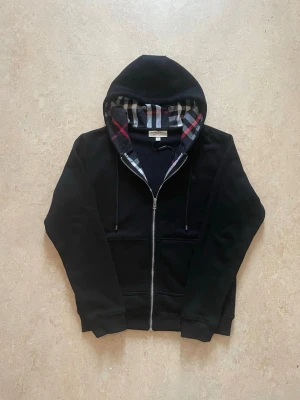 Svart hoodie med dragkedja Burberry - Snygg svart hoodie från Burberry med dragkedja och klassiskt rutigt foder i huvan. Mjuk bomullskänsla, två fickor fram och ribbade muddar. Perfekt för dig som gillar stilrena plagg med en twist av mönster. Storlek L men passar även om man har M.
