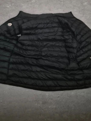 Svart väst från Moncler - Säljer en svart väst från Moncler med klassisk quiltad design och rundad krage. Västen har Monclers ikoniska logga på ärmen och ett färgglatt serietidningsfoder på insidan. MER INFO KOMMER PRIVAT.