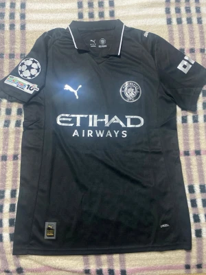 Svart Manchester City tröja Puma - Snygg svart Manchester City t-shirt från Puma med silverdetaljer och tryck. Tröjan har korta ärmar, klubbmärke och sponsorlogga framtill samt namn och nummer 10 'Cherki' på ryggen. Perfekt för fotbollsfans som vill sticka ut. Helt ny med tagg. Priset ej hugget i sten!