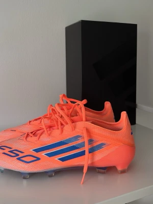 Adidas F50 CORAL BLAZE Laces Elite FG - Säljer ett par Adidas F50 fotbollsskor i en riktigt snygg orange färg med blå detaljer och klassiska tre ränder på sidan. Skorna har snörning och dobbar för spel på gräs. Lätta och smidiga i syntetmaterial, perfekta för snabba spelare som vill sticka ut på planen.
