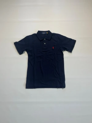 Mörkblå pikétröja från Ralph Lauren storlek S - Klassisk mörkblå pikétröja från Ralph Lauren med korta ärmar och krage. Tröjan har knappar framtill och den ikoniska röda loggan broderad på bröstet. Perfekt till jeans eller shorts för en stilren look. Den har storlek S, är helt ny och aldrig buren