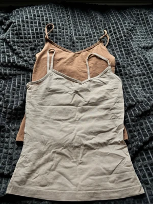 Två baslinnen i beige och brun - Säljer två enkla och stilrena linnen med smala axelband, ett i ljusbeige och ett i brunt. Båda har en rak passform och är tillverkade i mjuk bomull, perfekta att matcha till olika outfits.