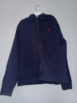 Mörkblå hoodie från Polo Ralph Lauren - Mörkblå hoodie med dragkedja från Polo Ralph Lauren. Klassisk design med röd broderad logga på bröstet, känguruficka och huva. Tillverkad i mjukt bomullsmaterial som är skönt att ha på sig. Perfekt för en avslappnad och stilren look.