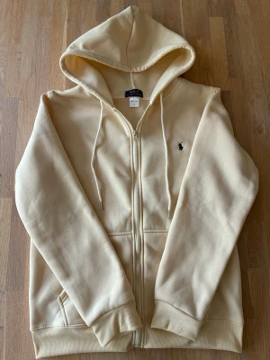 Ralph Lauren Zip hoodie beige - Snygg beige hoodie från Polo Ralph Lauren med dragkedja framtill och klassisk huva med dragsko. Broderad logga på bröstet och ribbade muddar vid ärmslut och nederkant. Perfekt för en chill och stilren look.