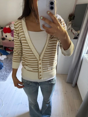 Beige och vit randig kofta 365 Sunshine - Snygg beige och vit randig kofta från 365 Sunshine. Koftan har v-ringning, knappar framtill och ribbade muddar. Perfekt att slänga över ett linne eller t-shirt för en avslappnad look. Sitter mer som en S.