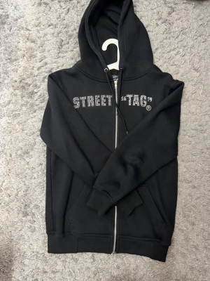 Svart hoodie STREET TAG by VIGGA - Svart hoodie med dragkedja från STREET TAG by VIGGA. Tröjan har huva med dragkedja och fickor framtill. Texten 'STREET TAG' i silvriga rine stones över bröstet ger en cool streetkänsla. Perfekt för en avslappnad och trendig stil.