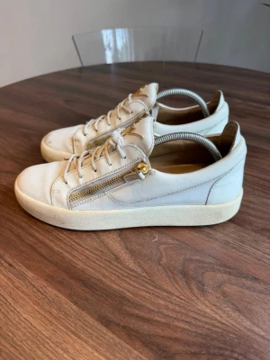 Vita sneakers från Giuseppe Zanotti - Snygga vita sneakers i läder från Giuseppe Zanotti med guldfärgade dragkedjor på sidorna och guldfärgad logga på plösen. Klassisk låg modell med vita snören och robust sula. Perfekta för dig som gillar exklusiva detaljer och streetstyle.