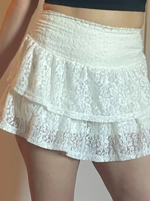Vit volangkjol i spets - Supersöt vit kort kjol med dubbla volanger och spetsdetaljer. Kjolen har shorts under och sitter som en smäck 💕har aldrig använts!