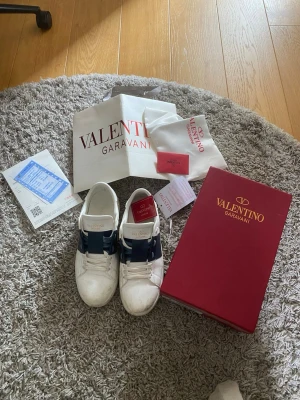Valentino Garavani vita sneakers - Snygga vita sneakers från Valentino Garavani med bred mörkblå rem över sidan och guldfärgad logga på plösen. Skorna har klassisk låg modell, vita snören och är tillverkade i läder. Perfekta för dig som vill ha en stilren och lyxig look.
