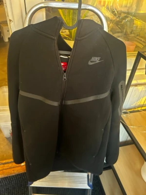 Svart Nike hoodie med dragkedja - Säljer en svart hoodie från Nike med hel dragkedja framtill och huva. Hoodien har diskreta grå detaljer över bröstet och ärmen samt Nike-logga på vänster sida. Tillverkad i mjukt material, perfekt för en sportig och avslappnad stil.