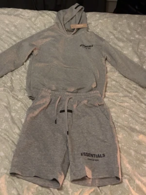 Grått set från Essentials Fear of God - Säljer ett grått set från Essentials Fear of God med hoodie och shorts. Setet är i mjukt bomullsmaterial och har tryckt logga på både tröjan och shortsen. Hoodien har dragsko och shortsen har resår i midjan med snörning. Hoodie + Shorts. Står L i kläderna men passar som M/S