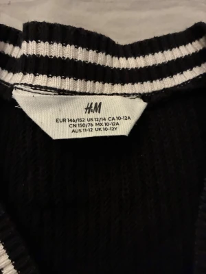 Svart stickad tröja med V-ringning H&M - Svart stickad tröja från H&M med vitrandiga detaljer runt V-ringad hals. Klassisk och enkel design med långa ärmar och ribbade muddar. Perfekt för lager på lager och passar till det mesta i garderoben.