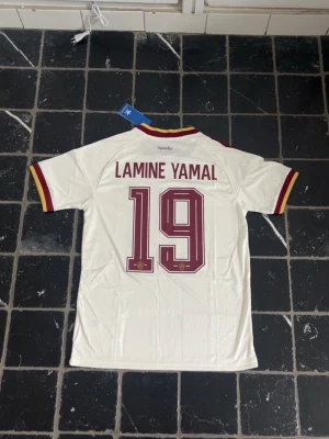 Spanien Lamine Yamal matchtröja - Vit Spanien fotbollströja från Adidas med Lamine Yamal och nummer 19 på ryggen. Tröjan har röda och gula detaljer på ärmslut och krage, samt röda Adidas-ränder på axlarna. Materialet är lätt och andas, perfekt för träning eller match.