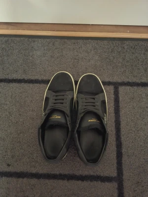 Svarta Saint Laurent sneakers - Svarta sneakers från Saint Laurent med vit sula och guldfärgad logga på sidan och plösen. Klassisk låg modell med snörning och ovandel i mocka och läder. Perfekta för dig som gillar stilrena och exklusiva skor. Tillkommer box, kort samt dustbag.