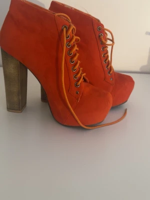 Orangea platåstövletter med snörning - Snygga stövletter i starkt orange mockaliknande material med höga, breda träfärgade klackar och platåsula. Skorna har snörning framtill med metallöljetter och en cool, edgy vibe. Perfekta för dig som vill sticka ut med din stil.