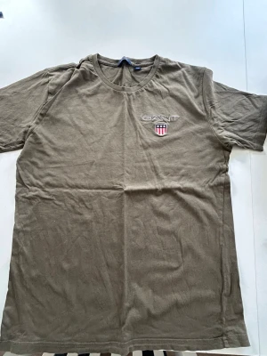Olivgrön GANT t-shirt med logga - Snygg olivgrön t-shirt från GANT med broderad logga och sköld på bröstet. Klassisk rund halsringning och korta ärmar. Tillverkad i mjuk bomull som känns skön mot huden. Perfekt för en avslappnad och stilren look.
