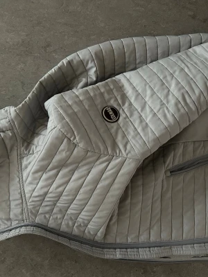 Ljusgrå pufferjacka från Colmar - Snygg ljusgrå pufferjacka från Colmar med quiltad design och diskret logotyp på bröstet. Jackan har dragkedja framtill och smala, horisontella sömmar som ger en modern look. Perfekt för höst och vår dagar som en sen sommarkväll och enkel att matcha med olika outfits.