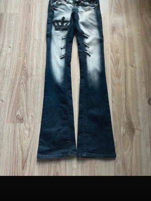 Bootcut jeans med krona och slitningar - Mörkblå bootcut jeans med ljusa slitningar och en stor svart krona som brodyr på vänster lår. Jeansen har fem fickor, låg midja och tydliga slitdetaljer framtill. Perfekta för en edgy streetstil. Innersöm: 21 cm Midja: 29 cm Längd: 99 cm