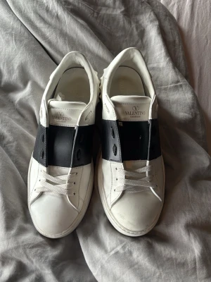 Valentino vita sneakers med svart band - Säljer ett par vita Valentino sneakers med bred svart läderdetalj över mitten och coola nitar runt hälen. Klassisk låg modell med vita skosnören och logga på plösen. Snygg och stilren design som sticker ut. De är lite smutsiga nu men innan jag skickar tvättar jag dem!